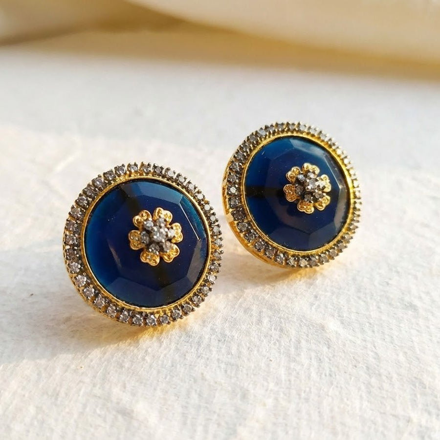 Close-up of navy blue round stud earrings with faceted stones surrounded by sparkling stones on light fabric