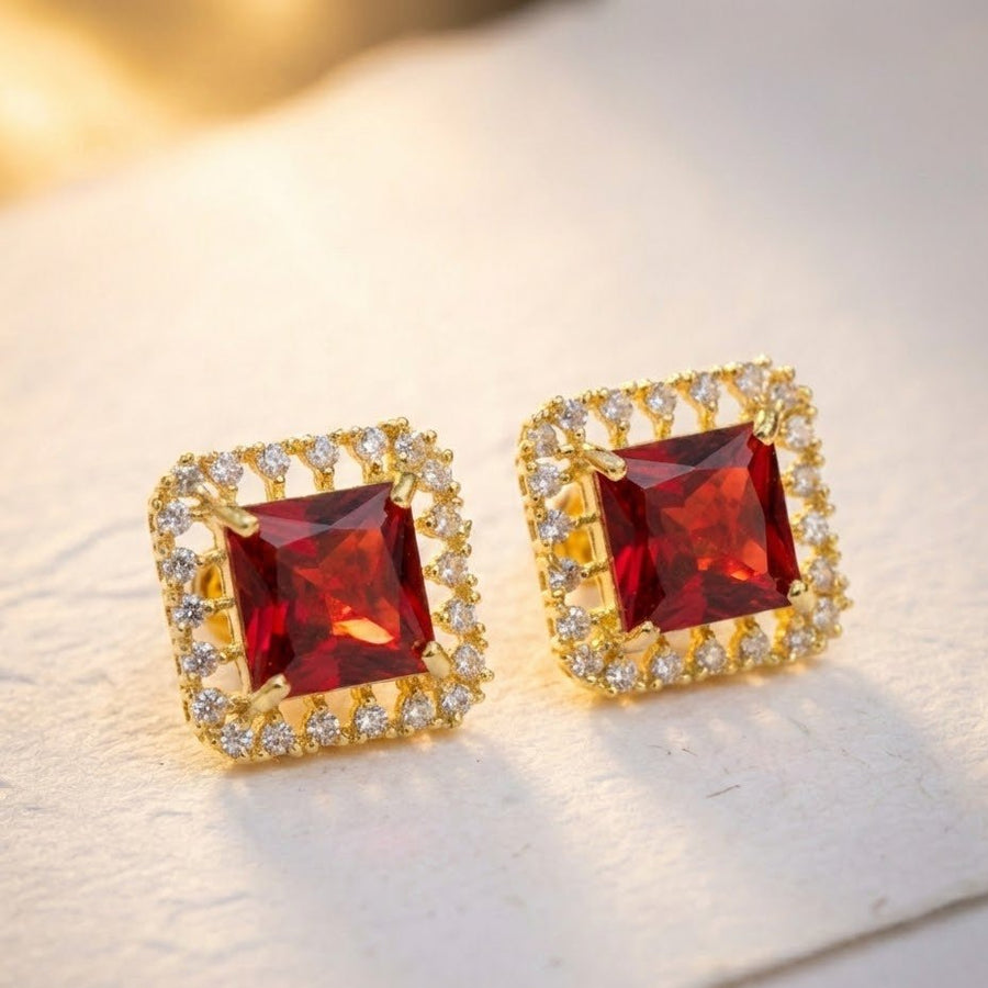 Lite ambient close-up of red square halo stud earrings on light surface highlighting stone clarity and gold-tone frame
