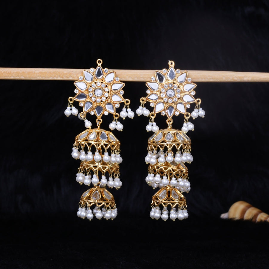 Pair of gold jhumka earrings with floral mirrored studs and triple-tiered pearl-adorned domes hanging from a wooden rod against black background