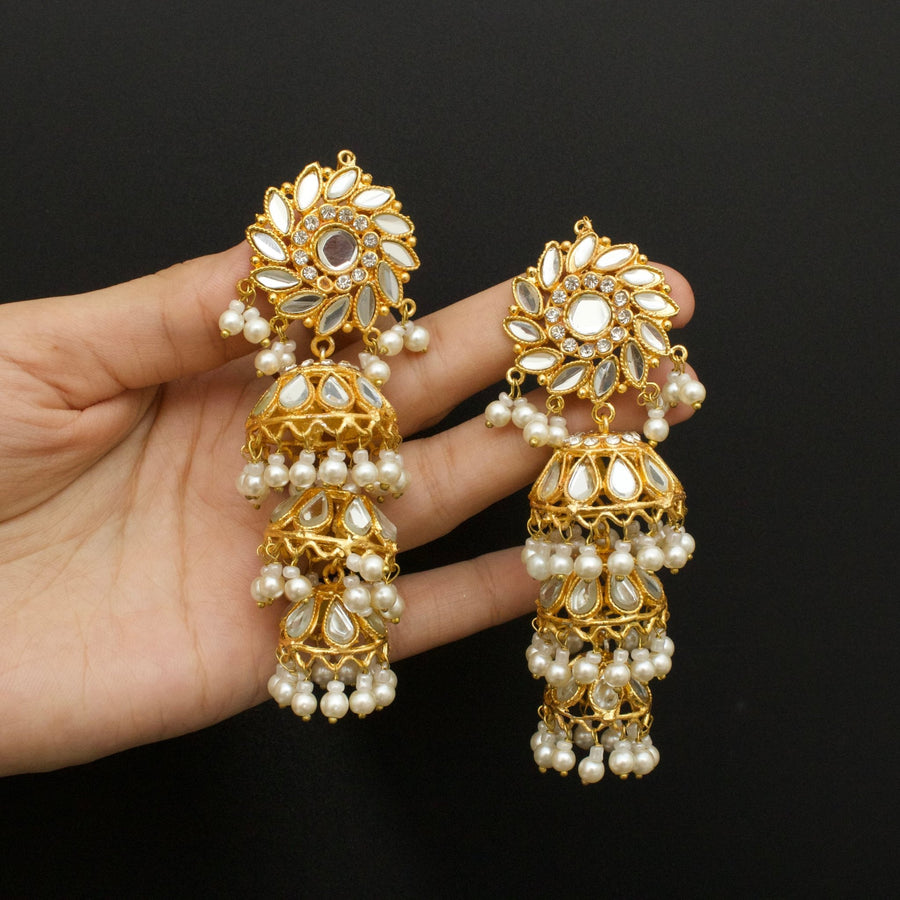 Hand holding a pair of gold-tone jhumka earrings embellished with white pearls and teardrop mirror stones on a black background