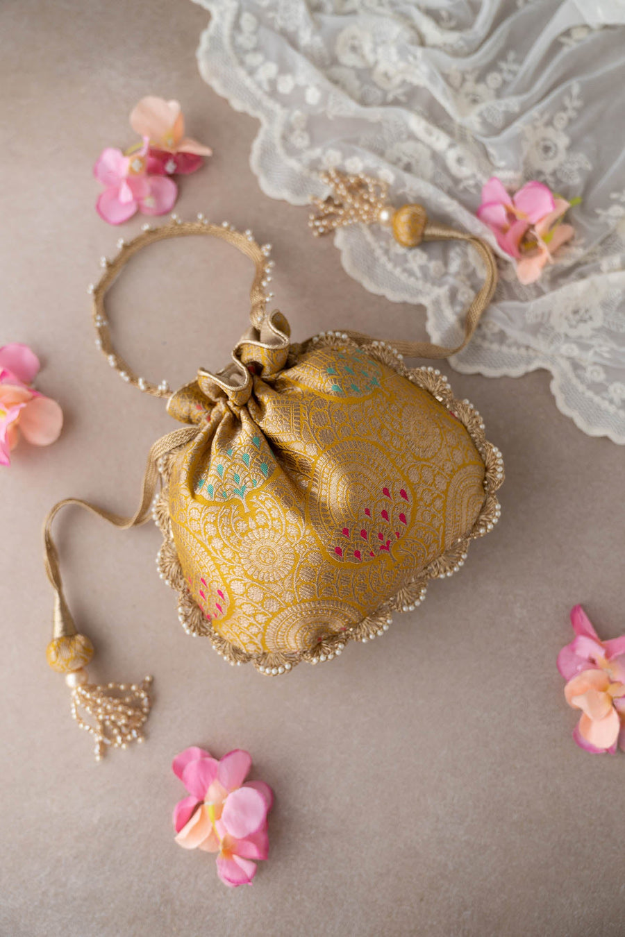 Elegant mustard gold Banarasi silk potli bag with floral brocade and pearl tassels displayed on a beige surface with lace and pink flowers