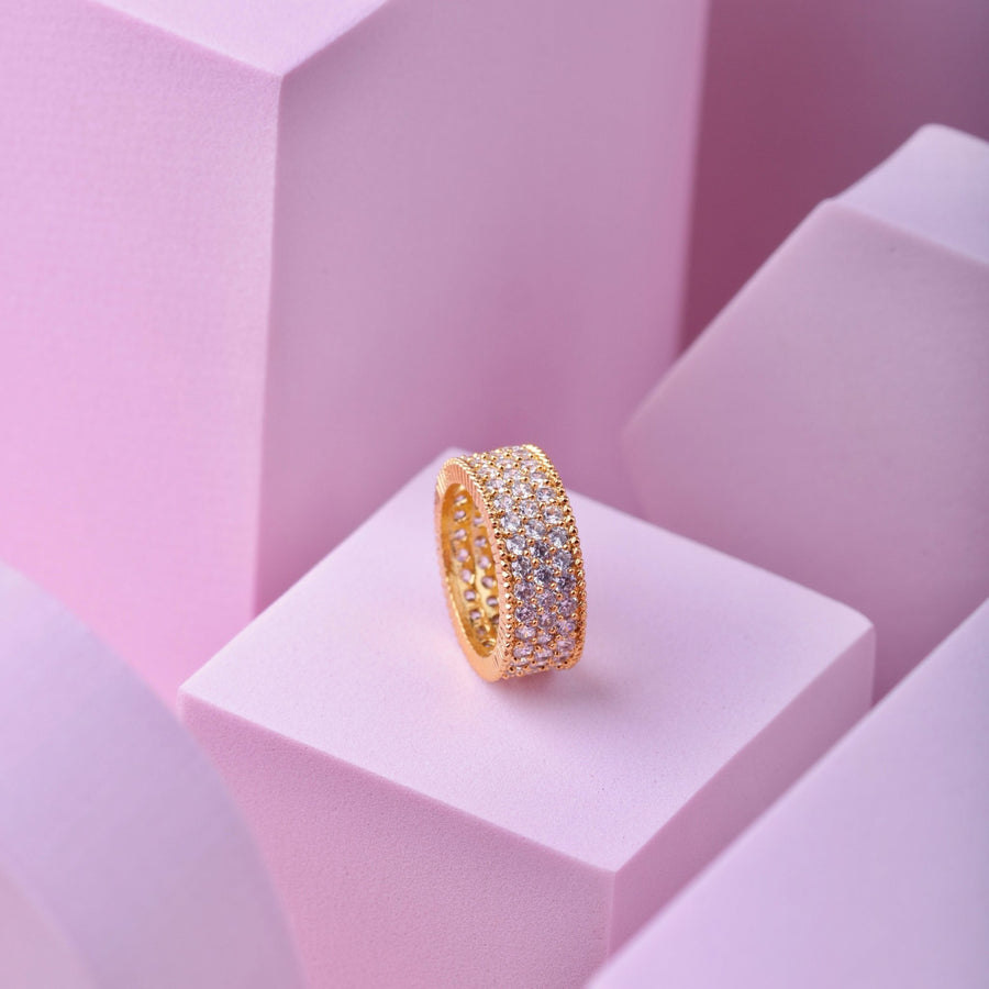 Gold-tone wide band ring completely encrusted with multiple rows of sparkling cubic zirconia stones on pink geometric display