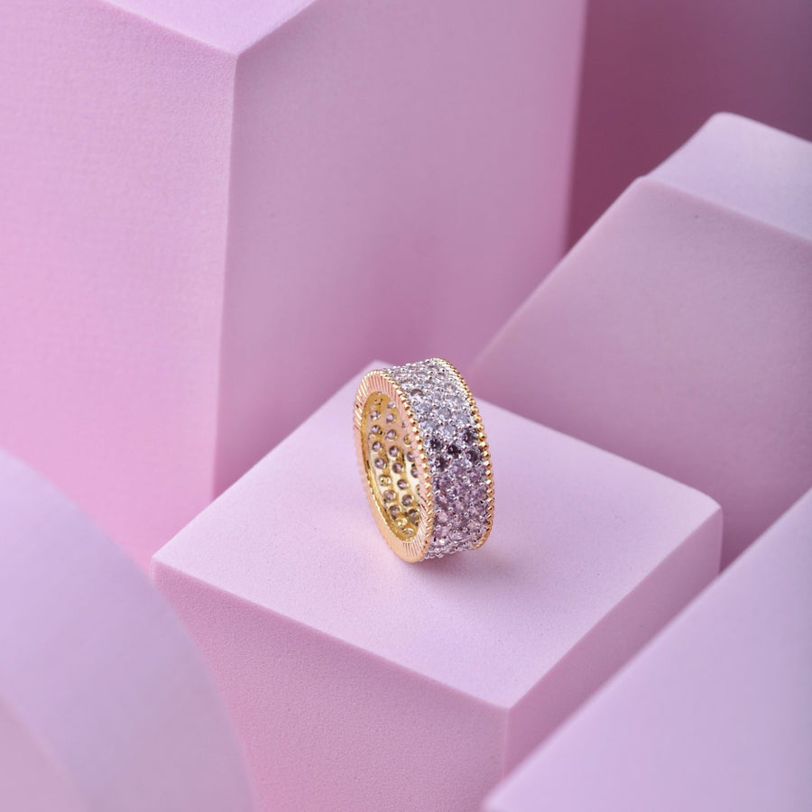 Close-up of gold-tone wide band cubic zirconia ring showing intricate milgrain edges on pink geometric block background