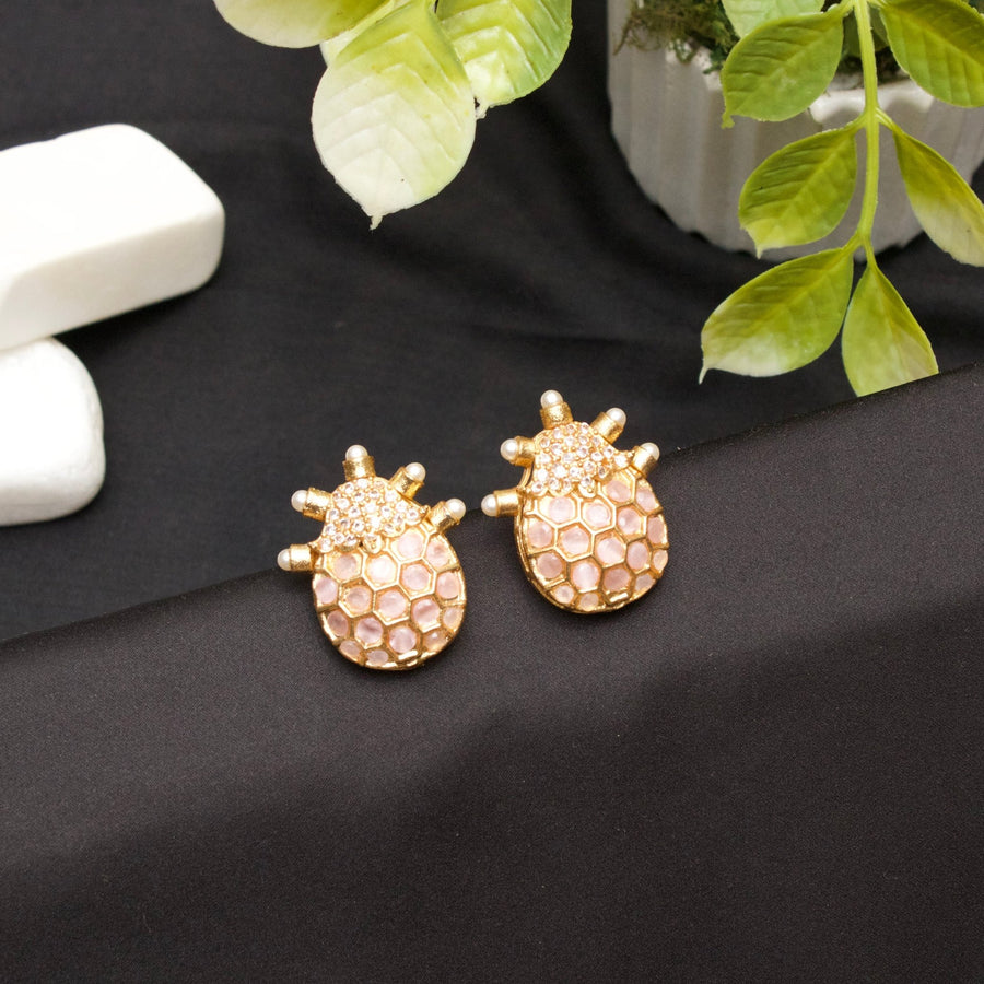 Pair of elegant gold-toned earrings with hexagonal pink stone design and pearl accent tips displayed on black fabric background with green plant leaves nearby